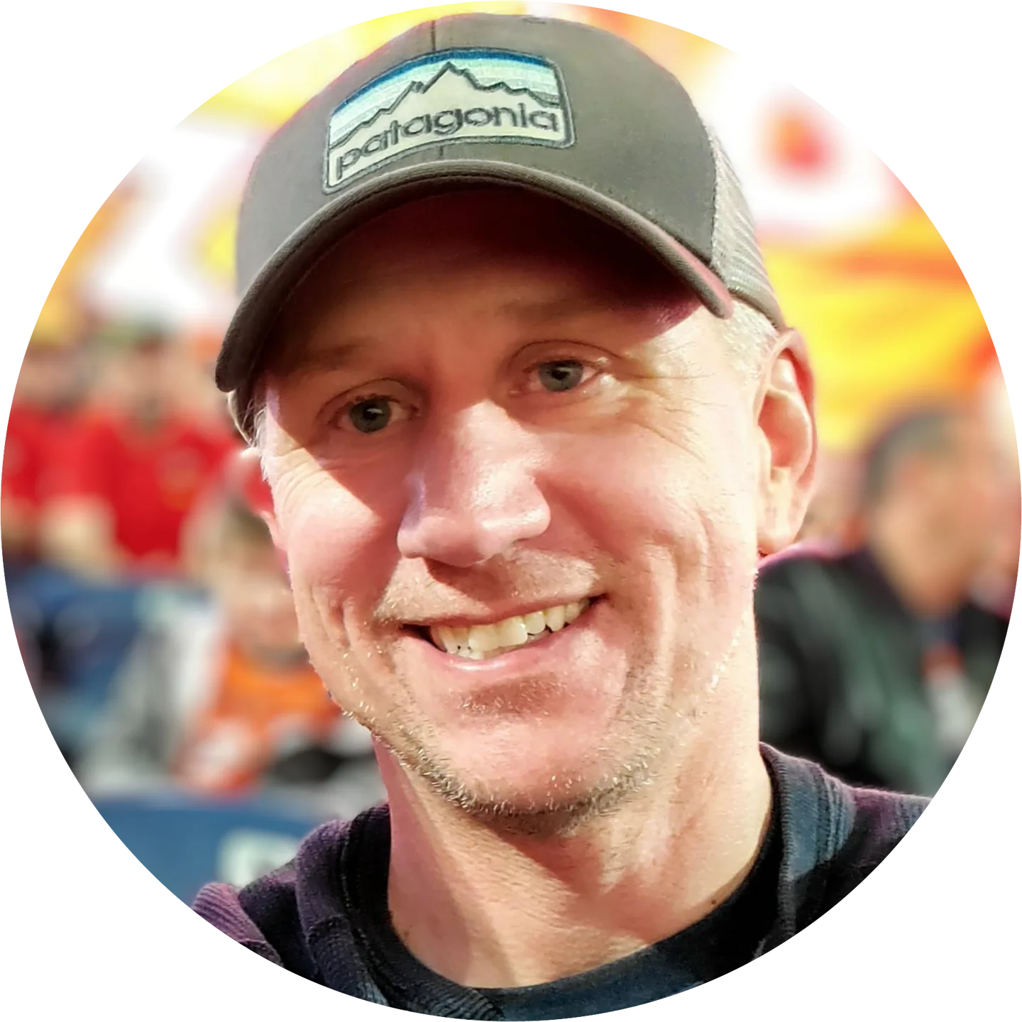 A smiling man wearing a gray Patagonia cap and a dark sweater, with a blurred background of people and orange tones.